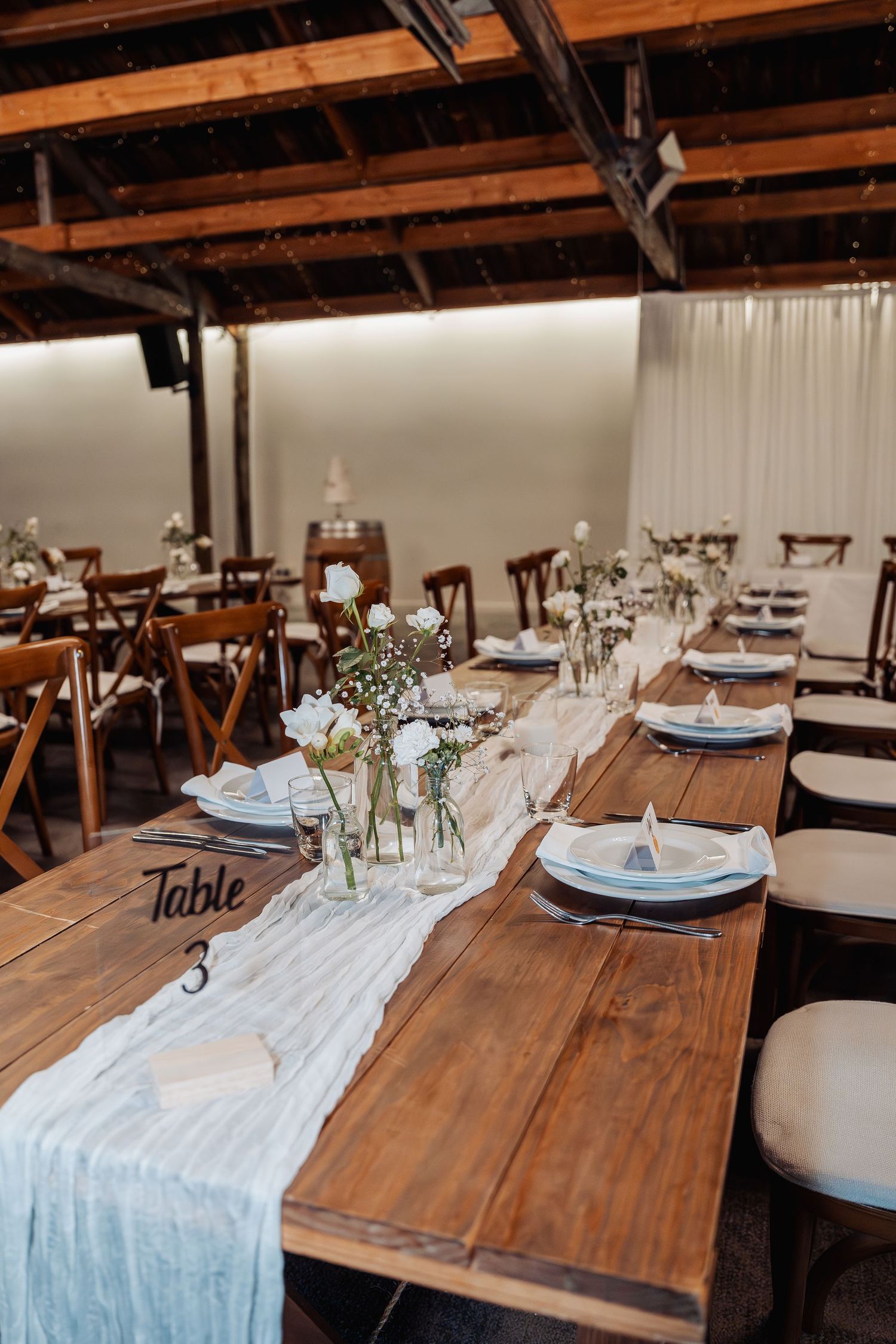 Rustic wedding reception table setup with blue runner, white floral centerpieces, and elegant place settings on wooden farm tables.