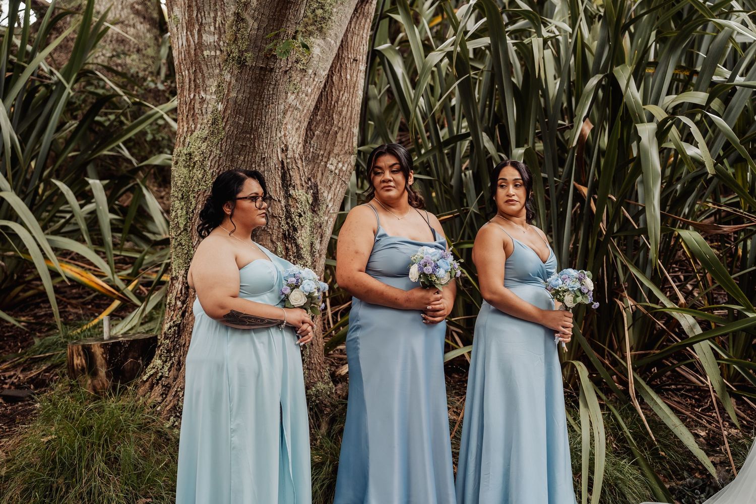 Three bridesmaids in light blue gowns holding bouquets stand together in a lush garden setting.