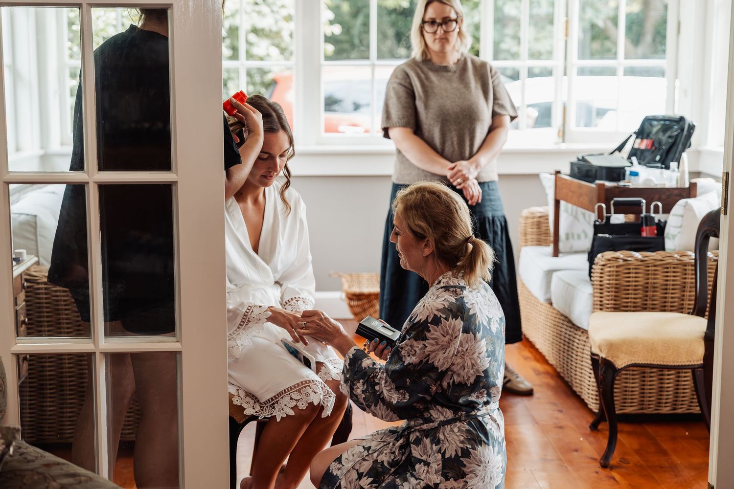 Bride in white robe getting ready as bridesmaid helps with accessories in a bright indoor setting.