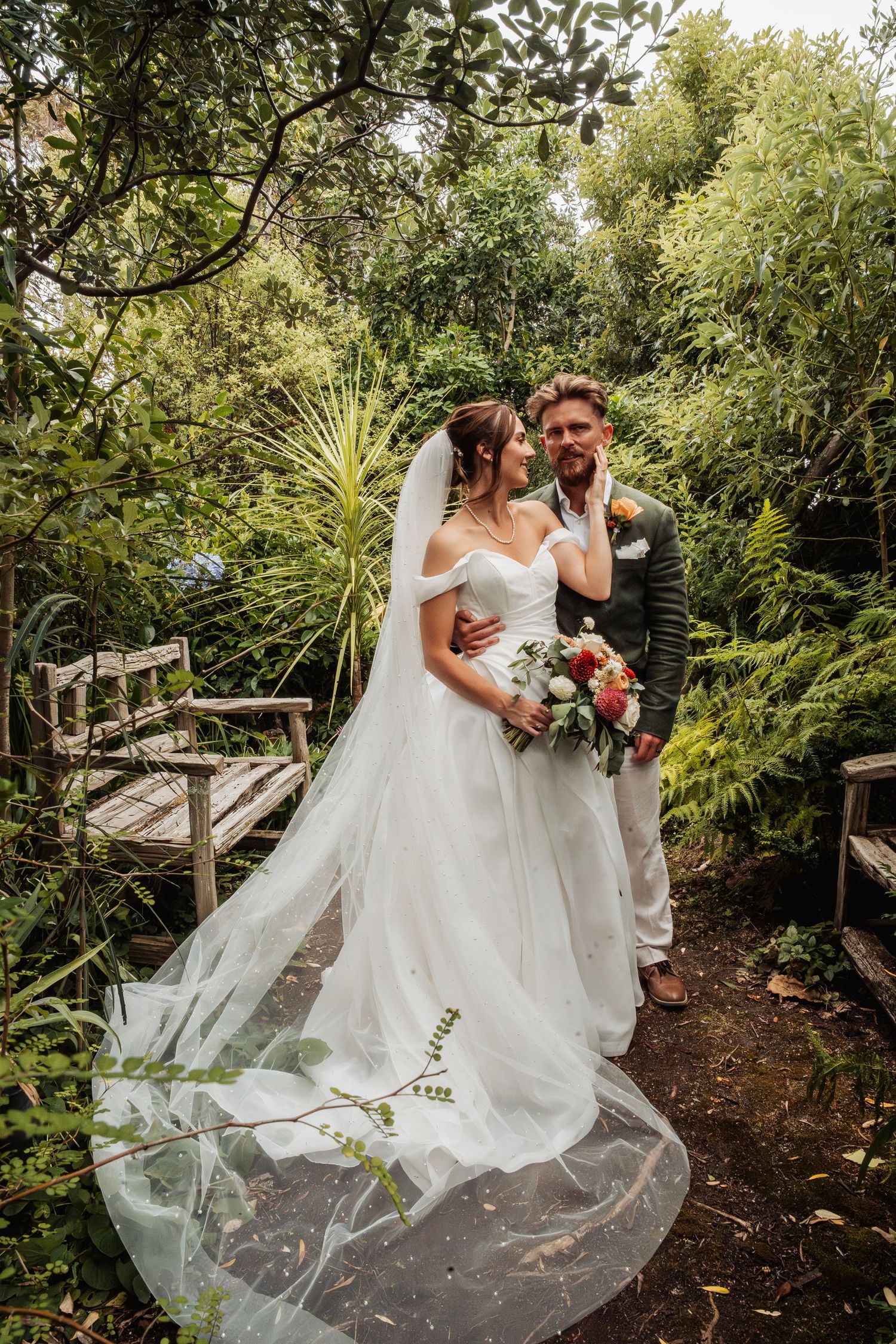 Bride and groom pose in lush green garden, bride wearing flowing veil and holding colorful bouquet.