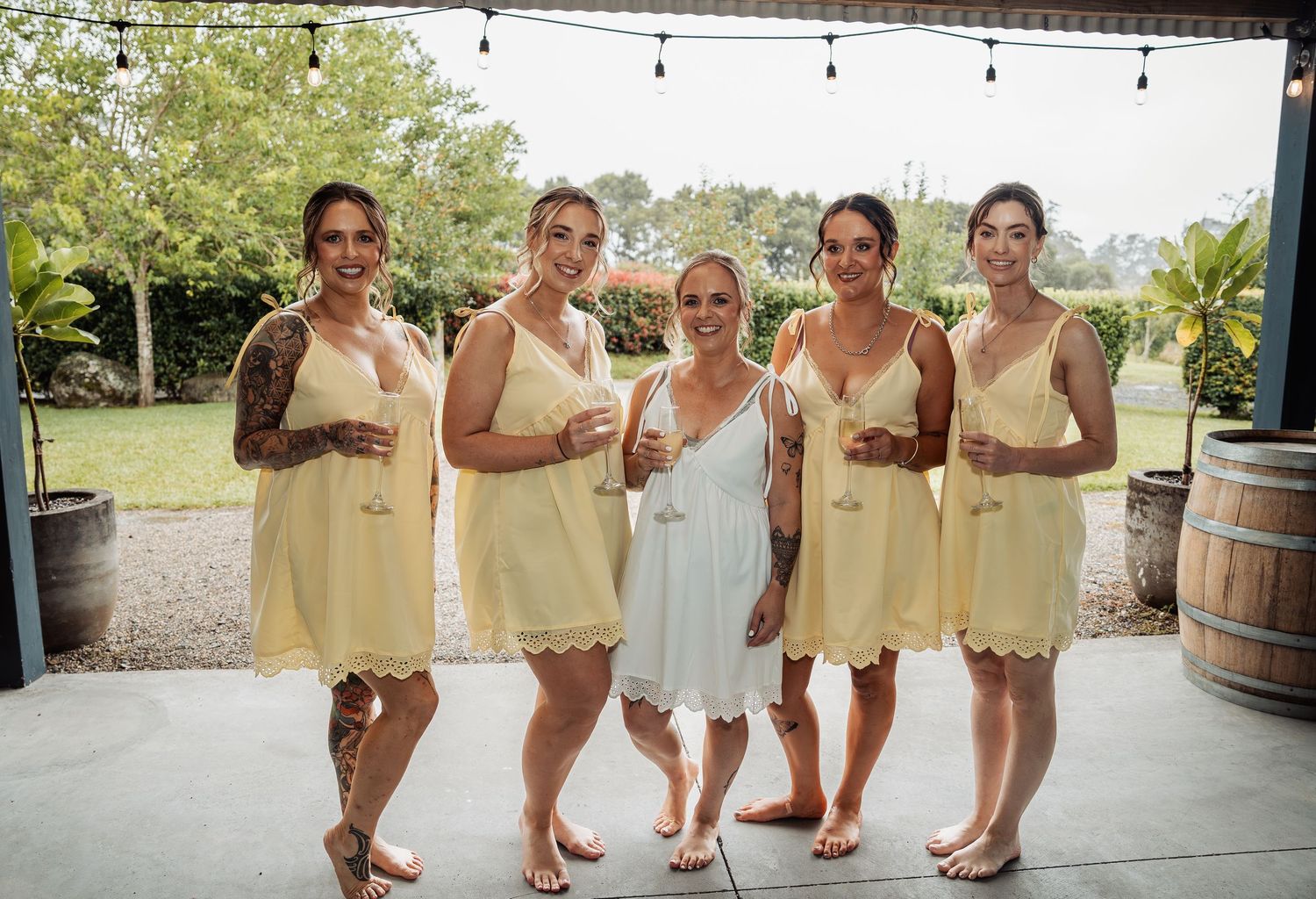 Bride in white dress poses with four bridesmaids in yellow dresses at an outdoor wedding venue.