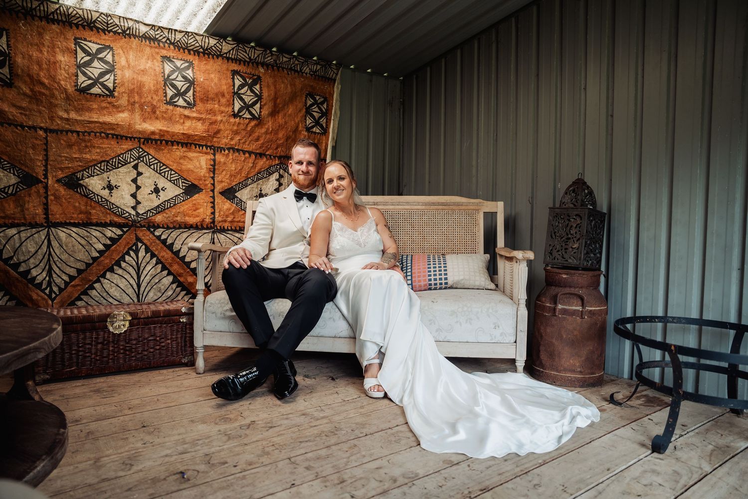 Bride and groom sitting together on a vintage sofa in front of a rustic tapestry backdrop on wooden floors.