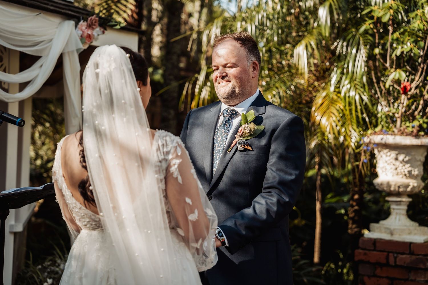 A romantic garden wedding ceremony shows a couple exchanging vows under lush tropical foliage and palm trees.