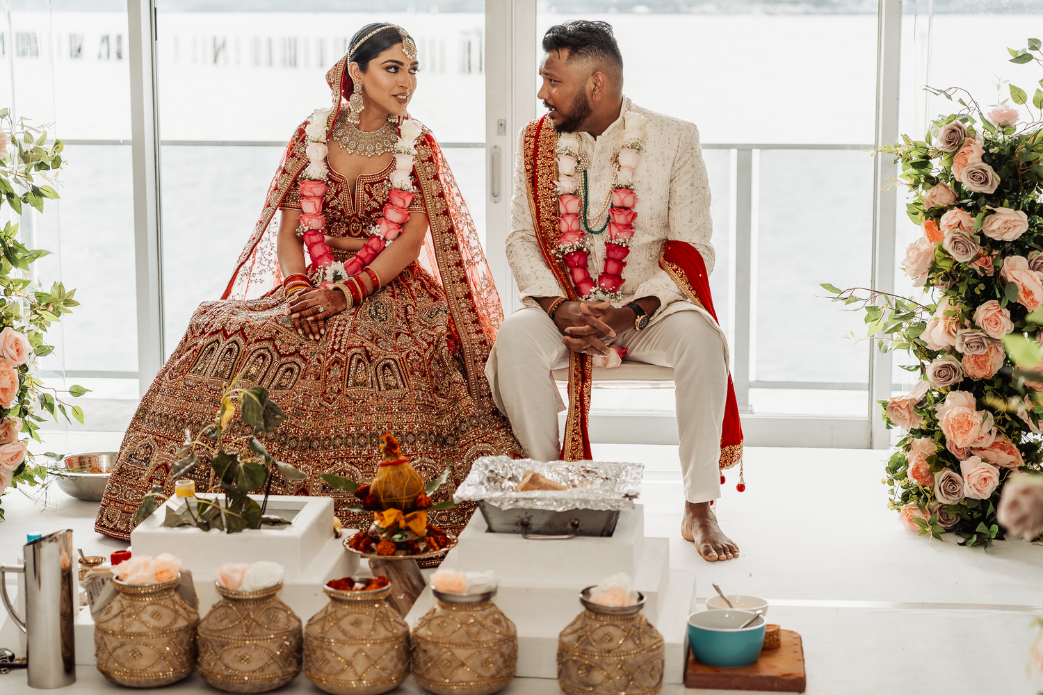 A couple sits together during their Indian wedding ceremony with traditional decorations and vessels.