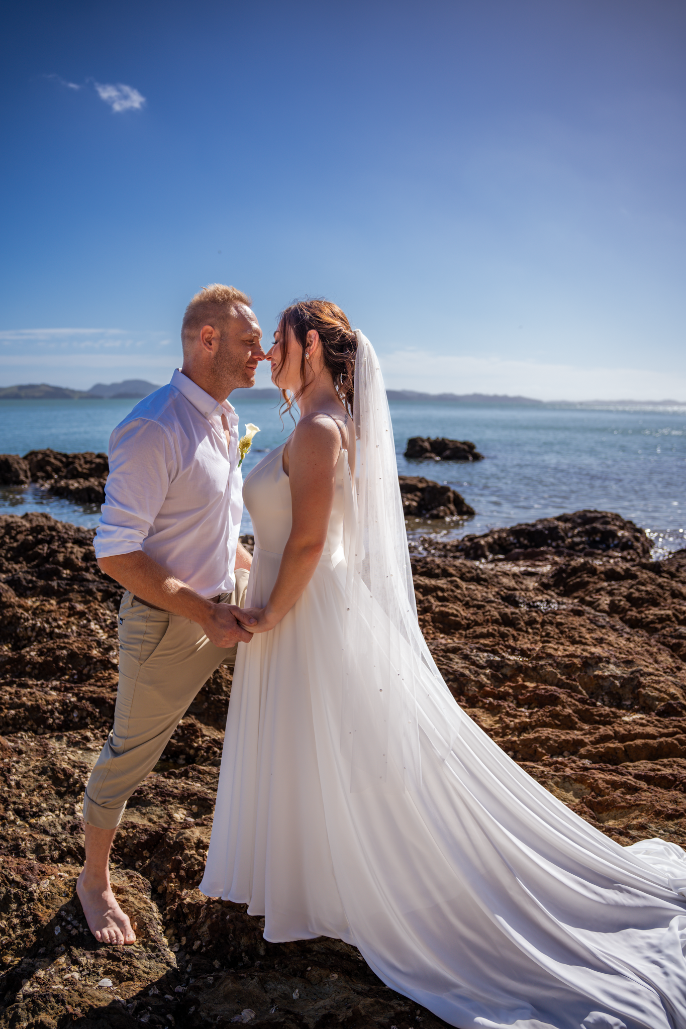 Romantic coastal wedding portrait with long white veil flowing against rocky shoreline.
