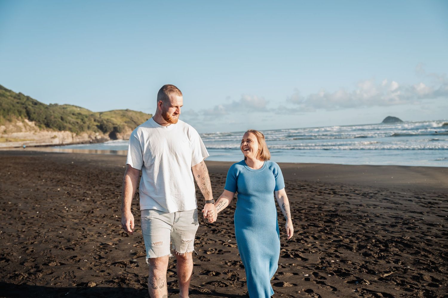 A couple walks hand in hand along a black sand beach with coastal cliffs and ocean views.