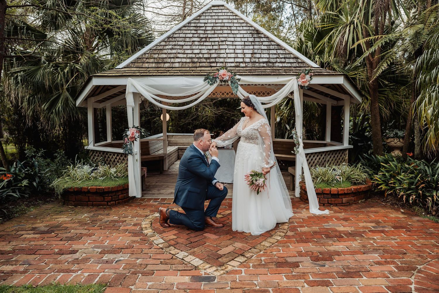 A couple shares a romantic moment on a rustic brick patio surrounded by lush tropical greenery and white gazebo.