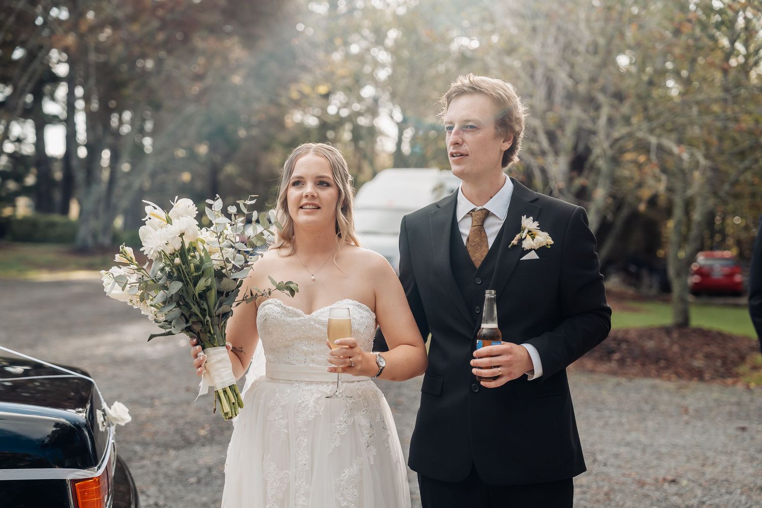 A joyful wedding couple walks outdoors in autumn, with the bride holding white flowers and groom holding a beer.