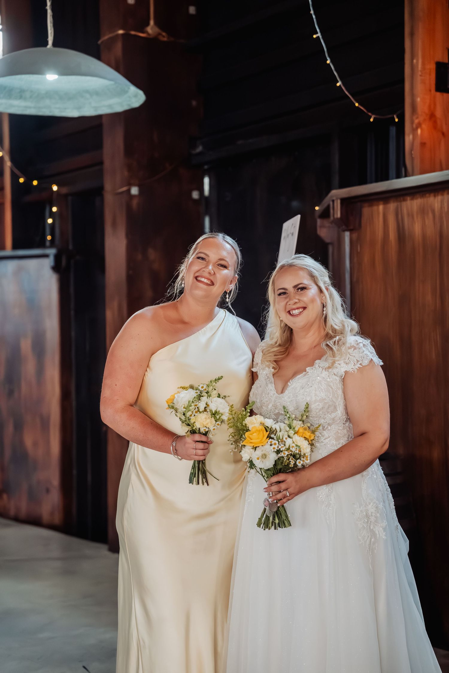 Two smiling brides in elegant white gowns holding floral bouquets stand together inside a warm barn setting.