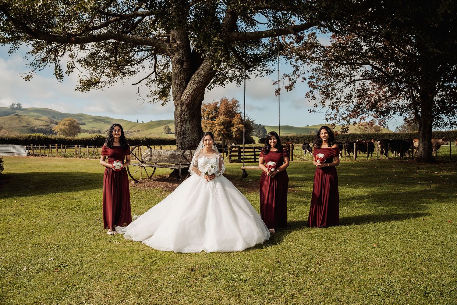 Bride in white ball gown poses with four bridesmaids in burgundy dresses on lush green farm grounds.