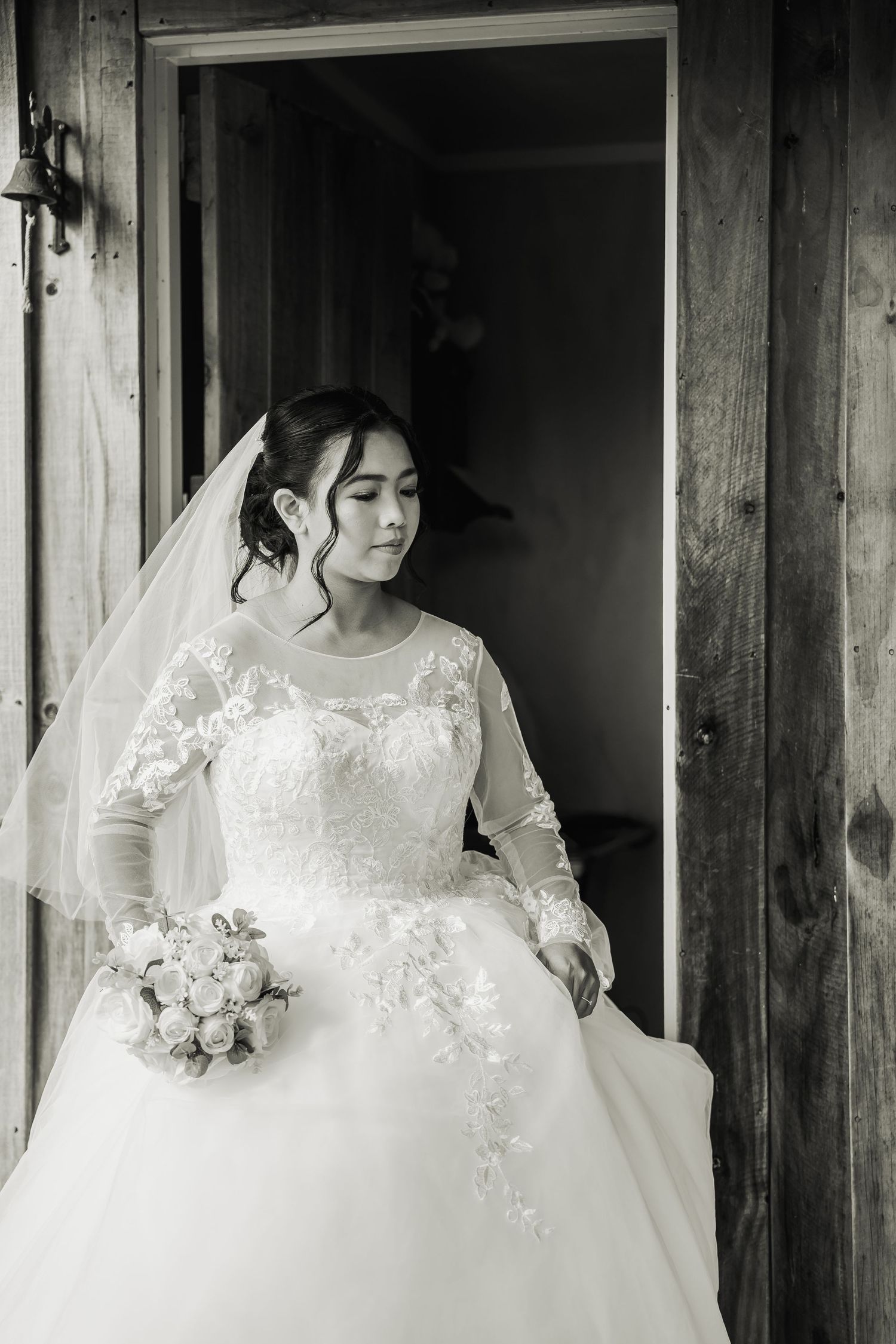 Bride in lace long-sleeve wedding gown holding bouquet, standing in doorway, black and white photo.