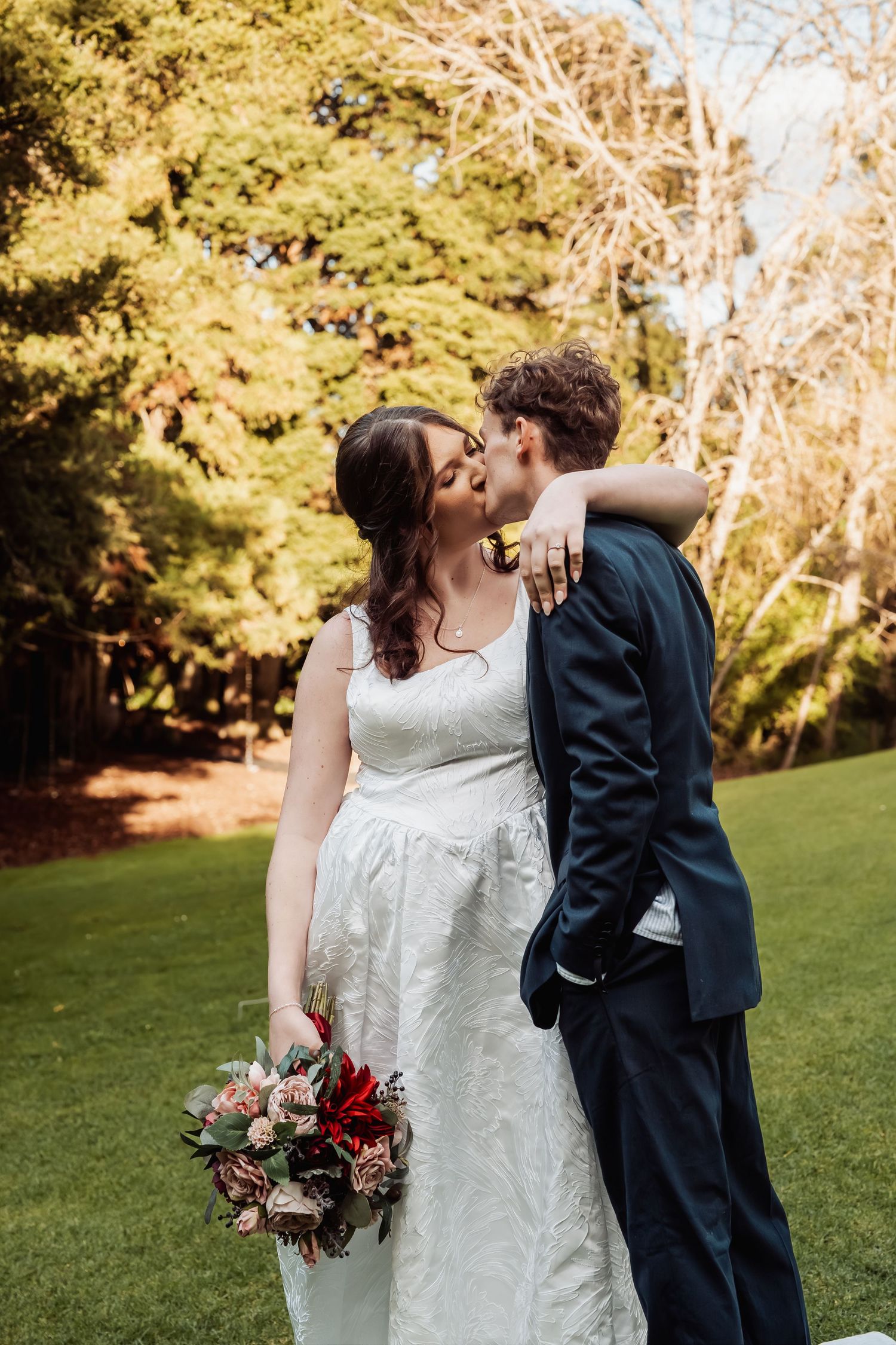 Bride and groom sharing a kiss outdoors on green lawn, bride holding red bouquet, groom in navy suit, lush trees background.