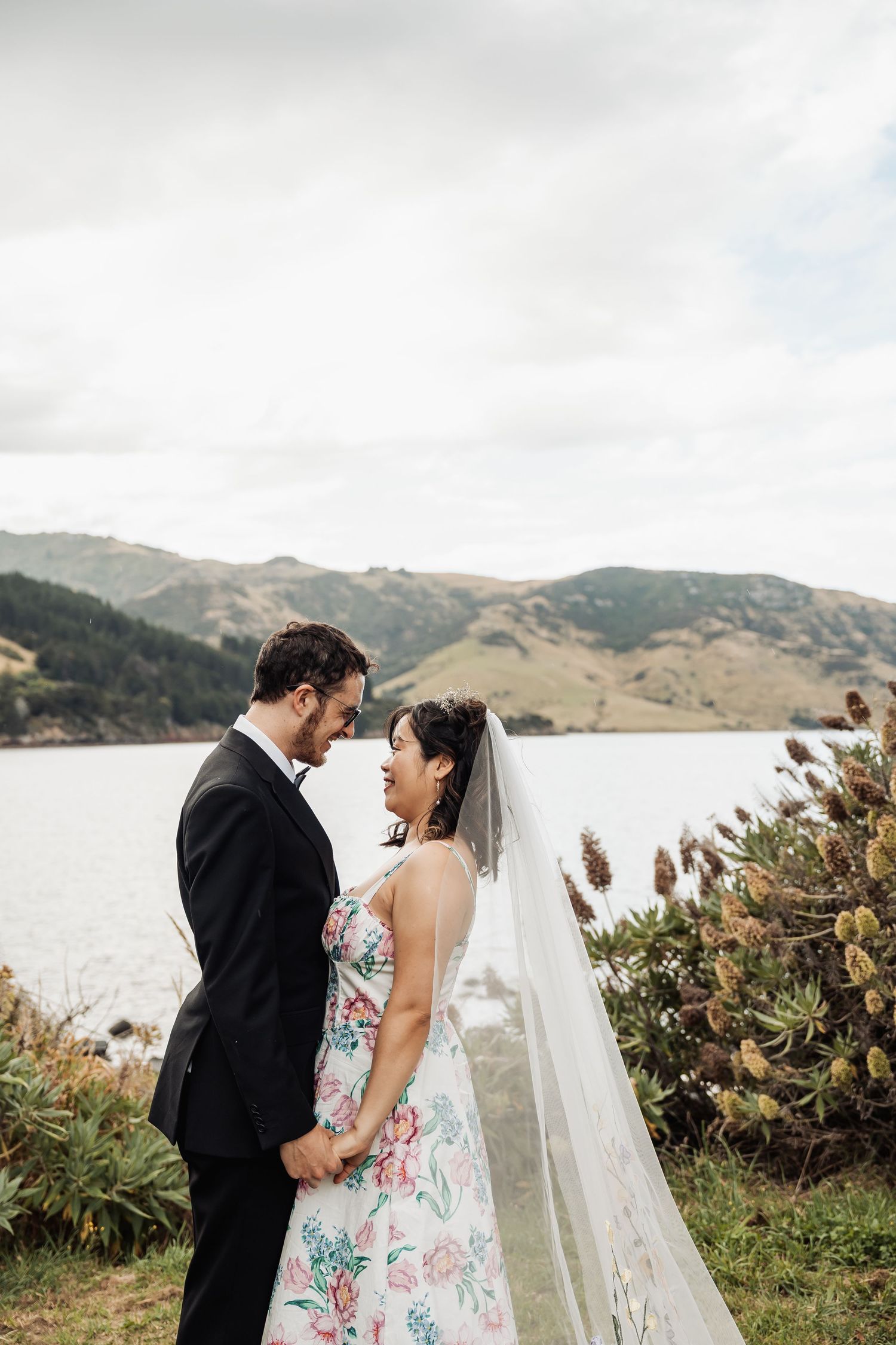 Bride and groom hold hands facing each other with scenic coastal hills and water in the background on their wedding day.
