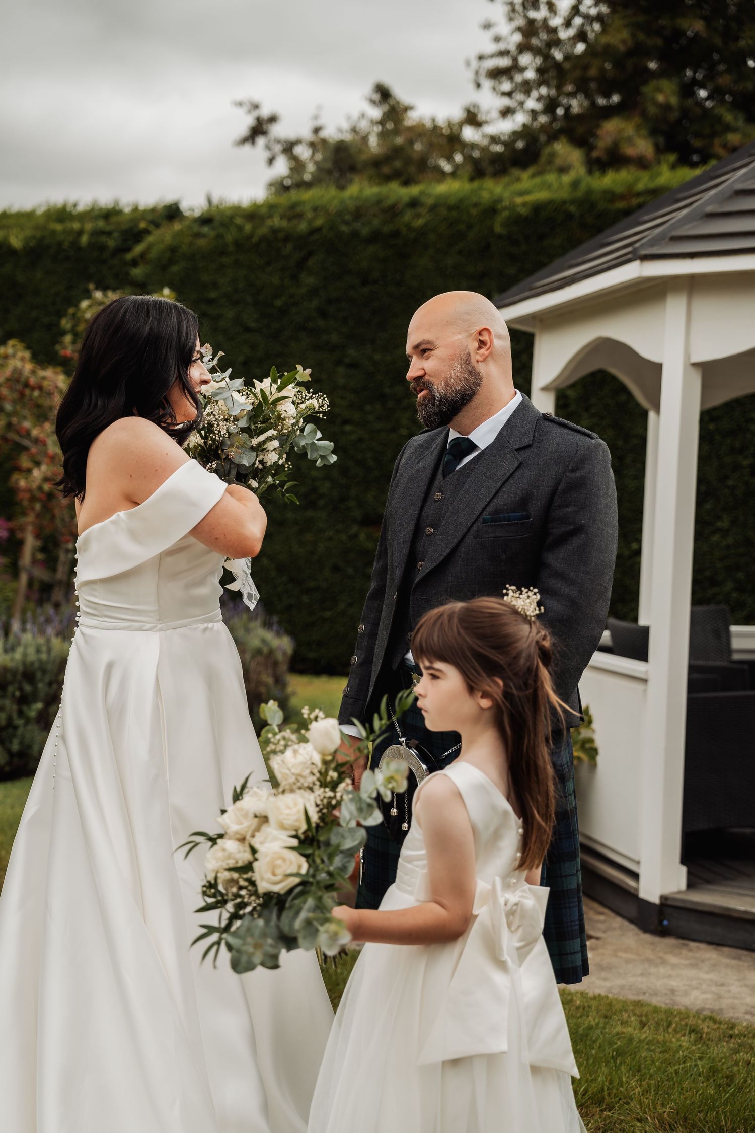 Bride in off-shoulder gown holds bouquet with bald groom in kilt and flower girl in white dress in garden.