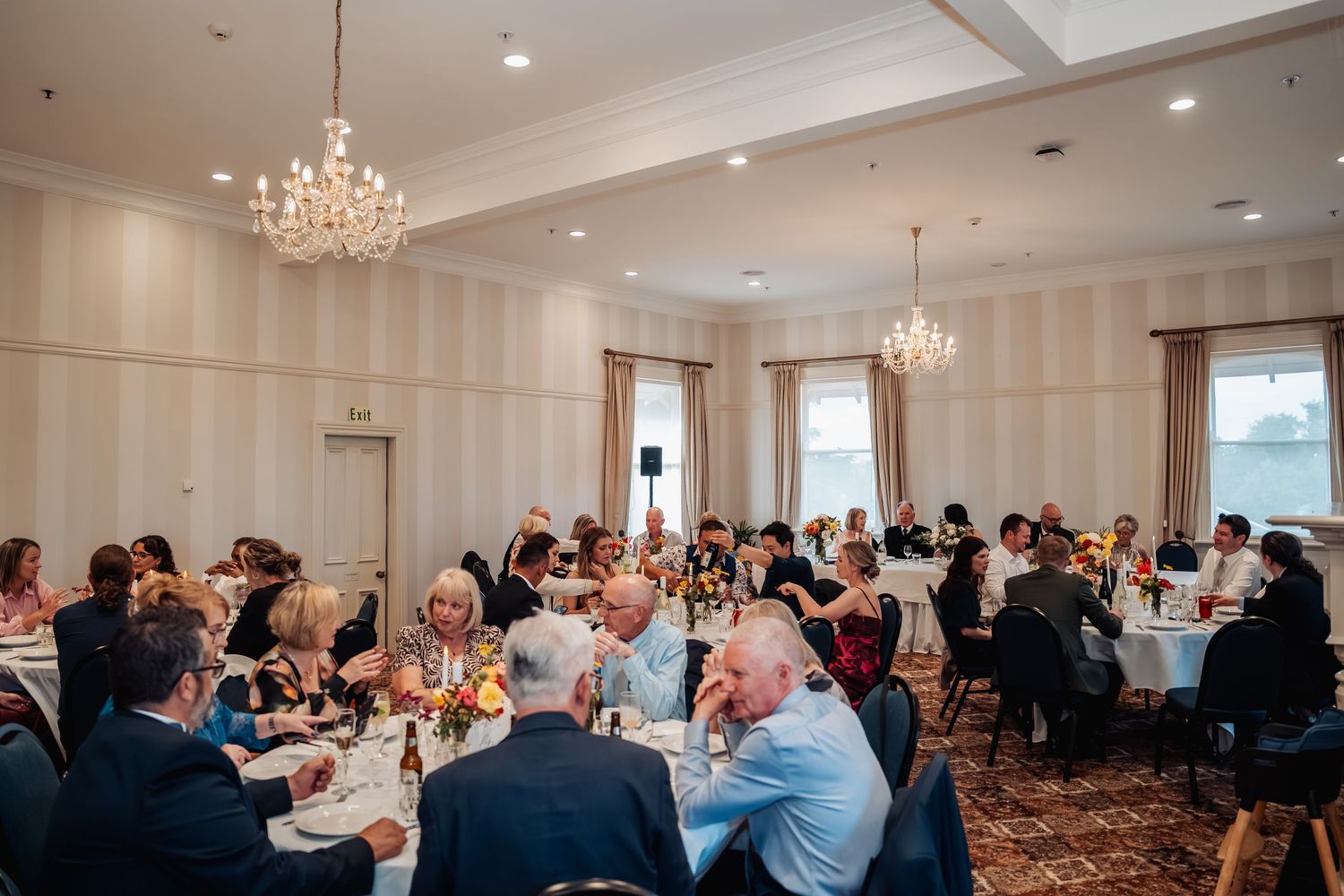 Elegant wedding reception in a chandelier-lit ballroom with guests seated at round tables adorned with floral centerpieces.