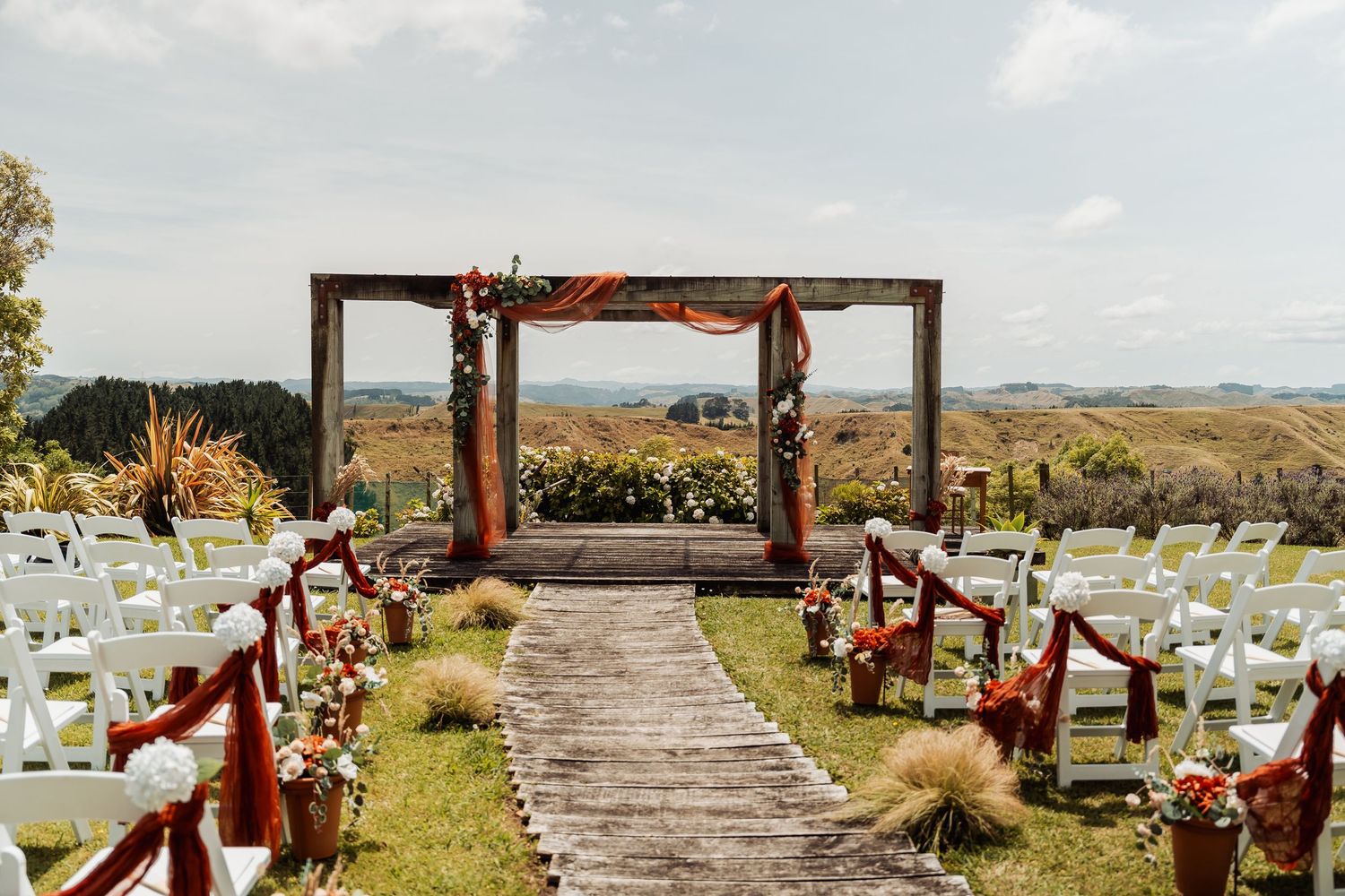Elegant outdoor wedding ceremony setup with wooden arch, floral decorations, and white chairs overlooking scenic hills.