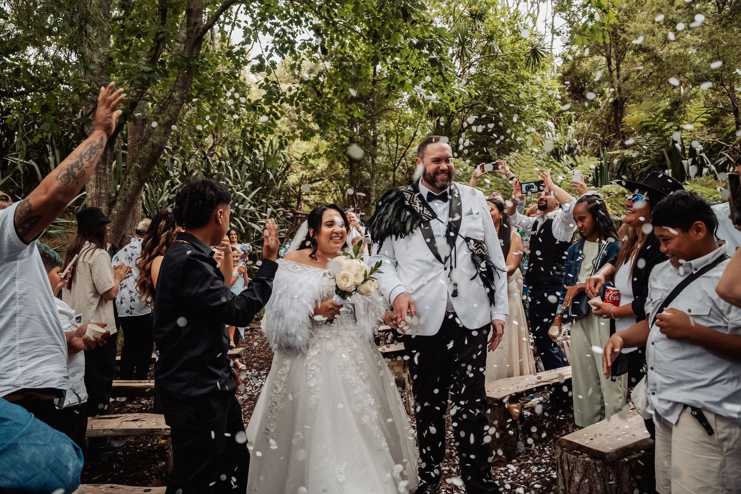 Joyful newlyweds walk through confetti shower among celebrating guests at outdoor woodland wedding.