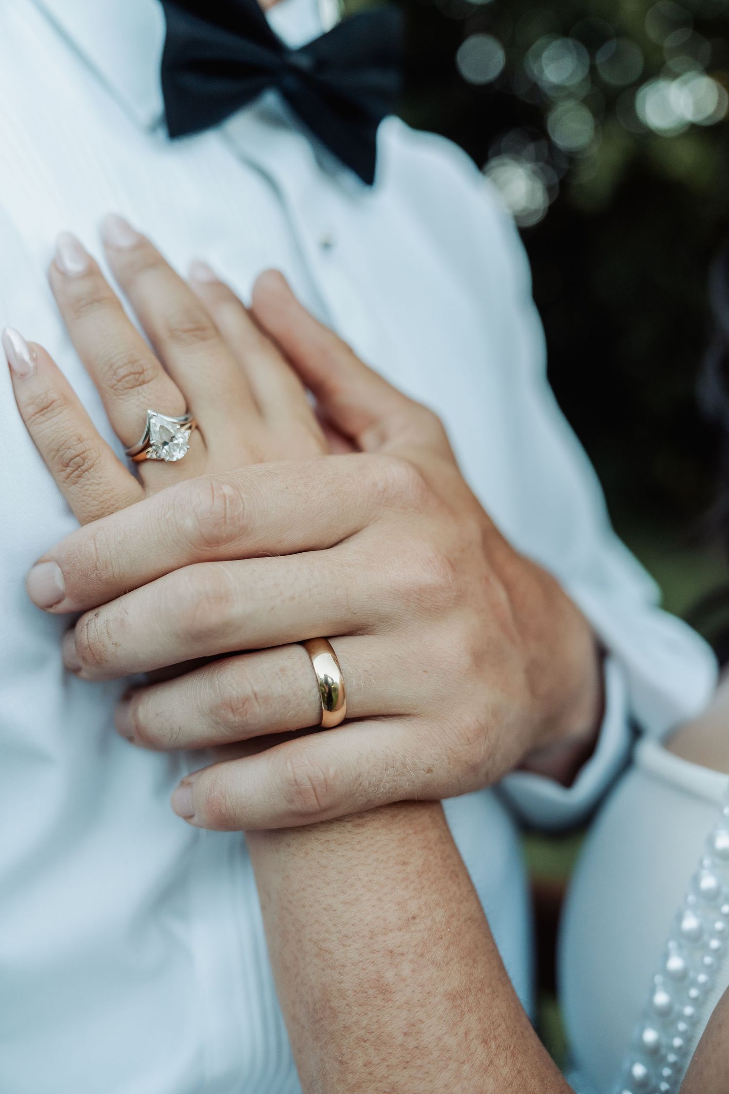 Close-up of newlyweds hands showing diamond engagement ring and gold wedding band against white shirt.