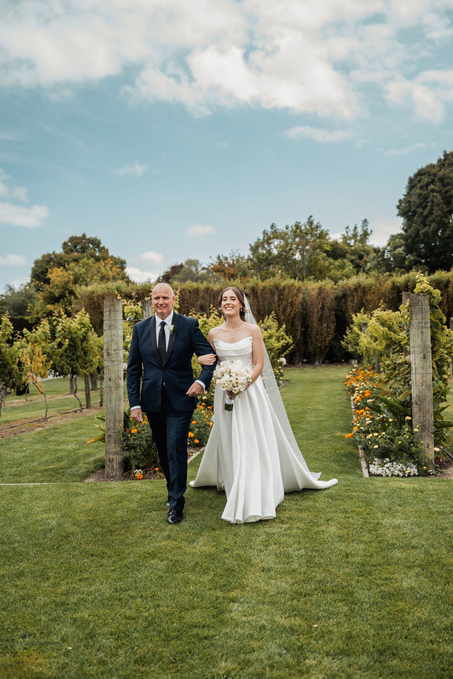 Bride in white gown walks arm-in-arm with suited man through lush garden vineyard.