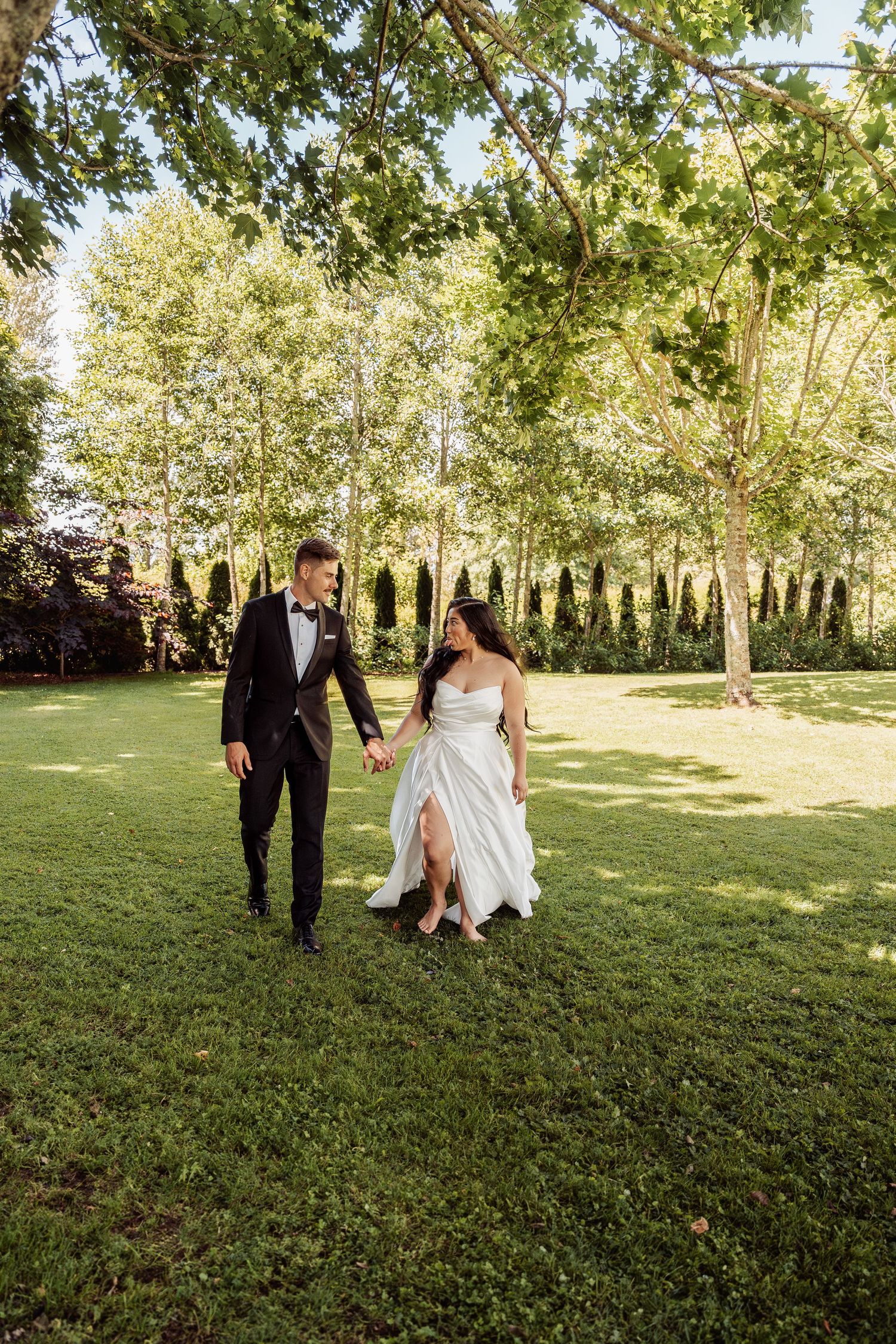 Bride and groom walk hand-in-hand across lush green lawn surrounded by tall trees on their wedding day.