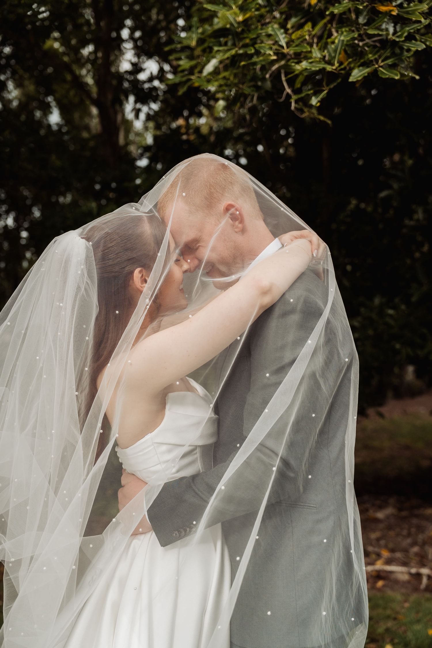 Bride and groom share an intimate moment hidden beneath a pearl-adorned wedding veil outdoors.