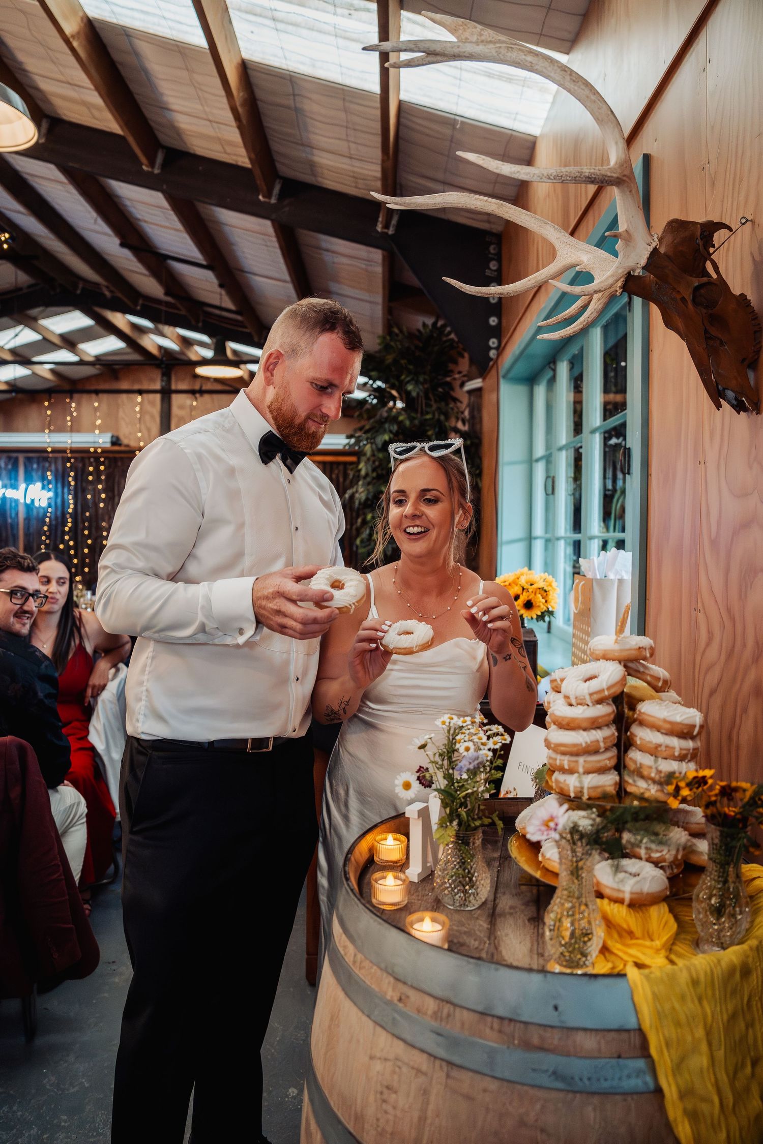 Bride and groom smile while selecting donuts from a rustic barrel dessert display at their wedding reception.
