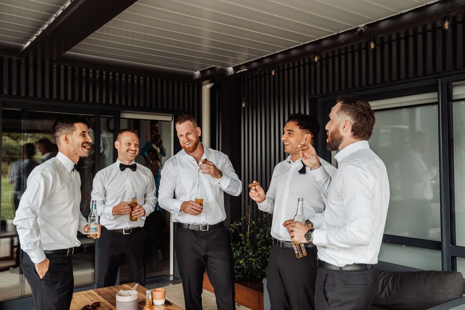 Five groomsmen in white shirts and black bow ties laughing and enjoying drinks together at a wedding celebration.
