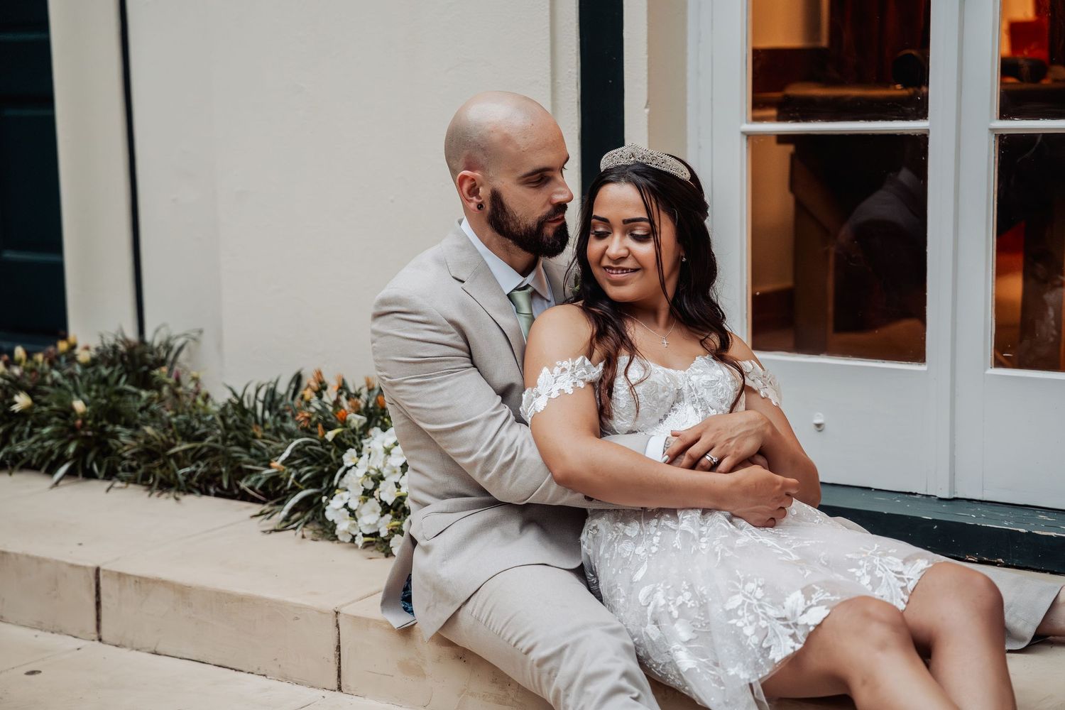 A bald groom in a beige suit tenderly embraces his bride in a lace dress on outdoor steps.