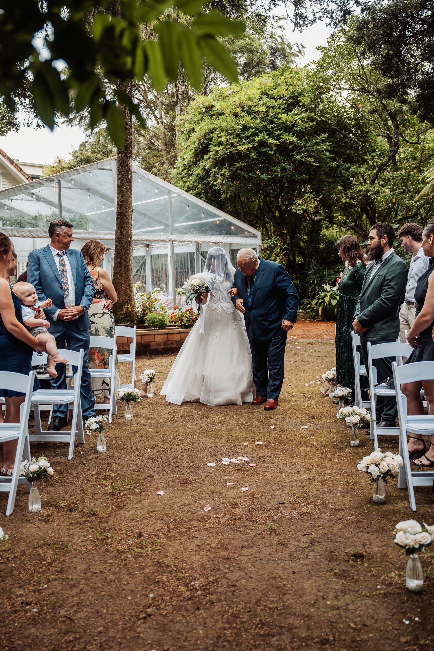 Bride walking down outdoor garden aisle with father during wedding ceremony with guests seated.