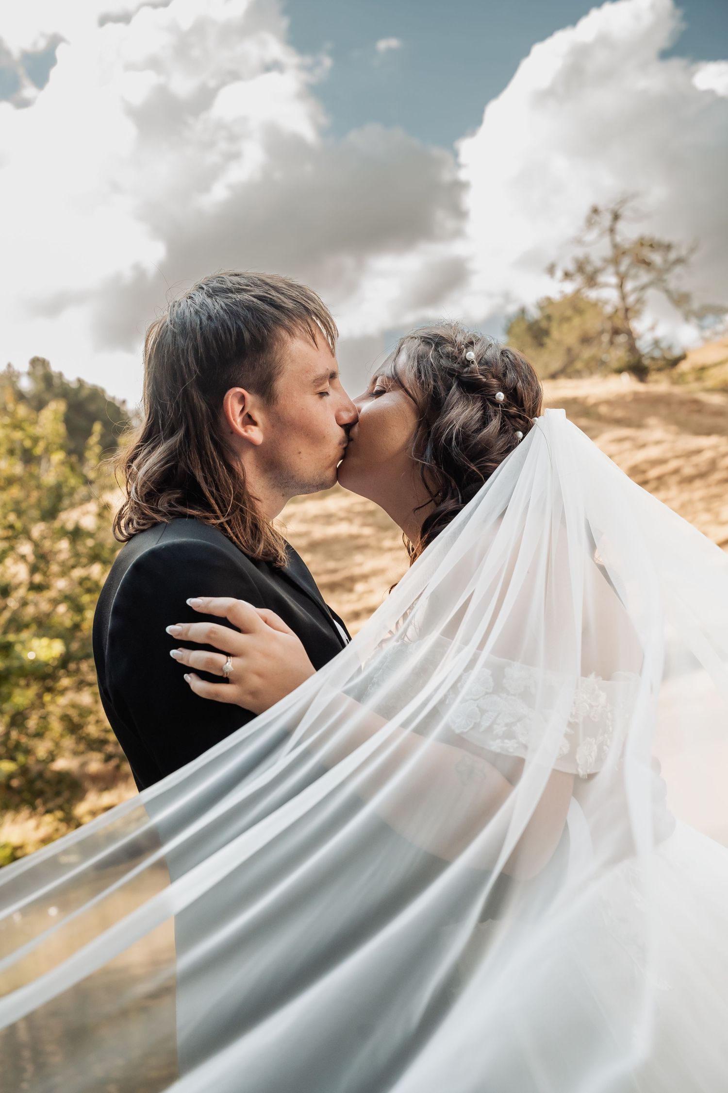 Bride and groom kissing under billowing white veil against dramatic cloudy blue sky backdrop.