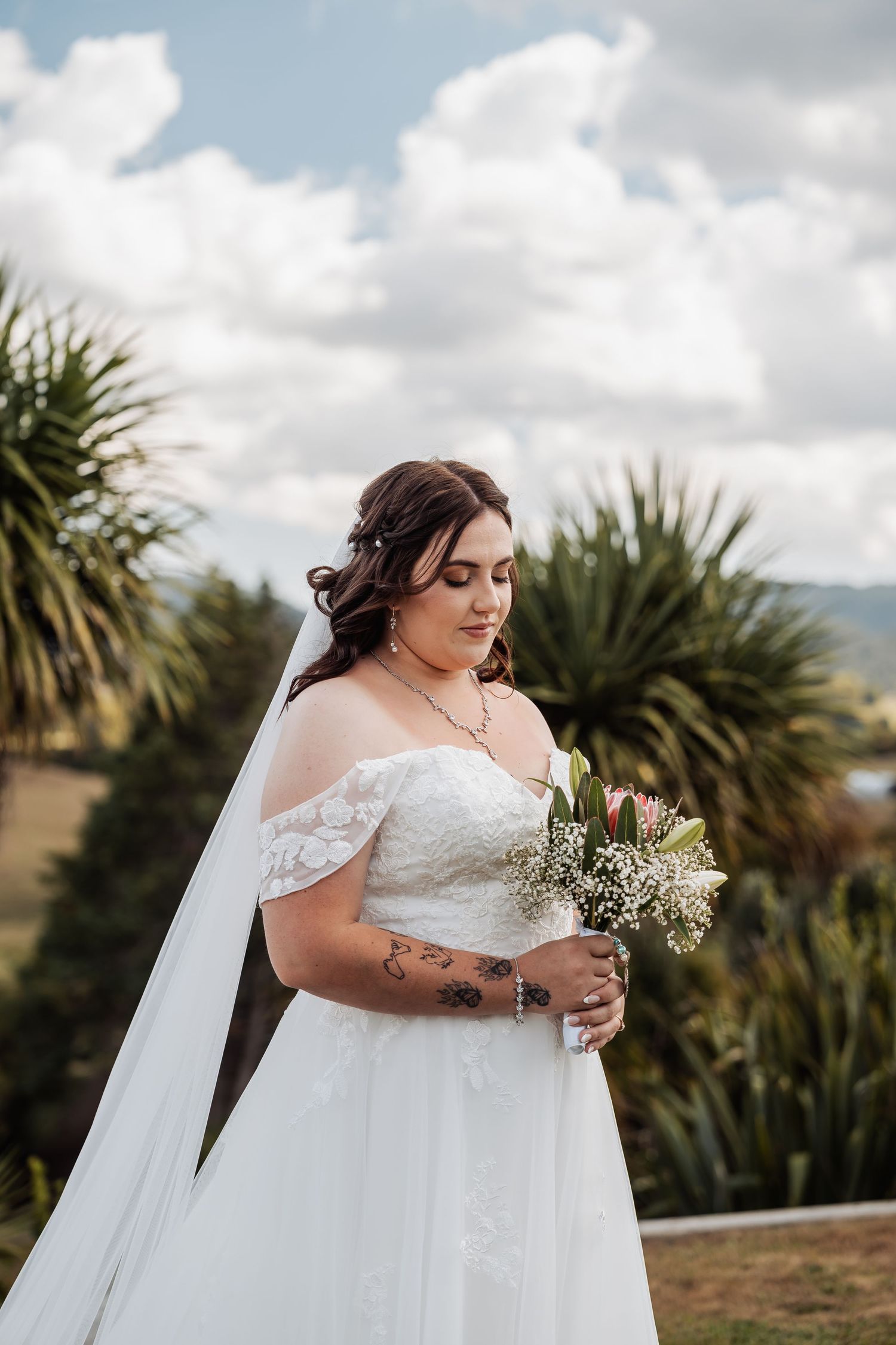 Beautiful bride in lace off-shoulder gown with veil looks down at bouquet with palm trees behind her.