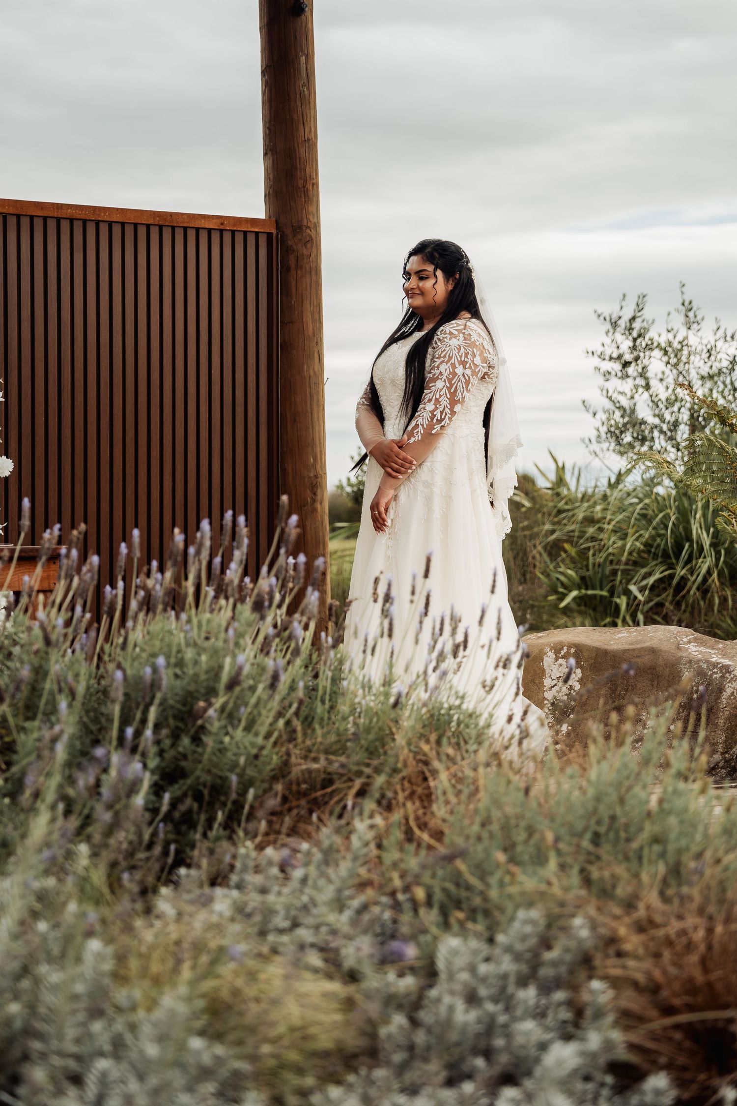 Bride in white lace wedding dress stands among lavender plants beside rustic wooden fence outdoors.