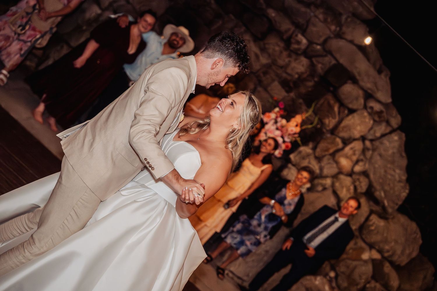 Groom dips bride during romantic first dance inside a warmly lit stone-walled reception venue.