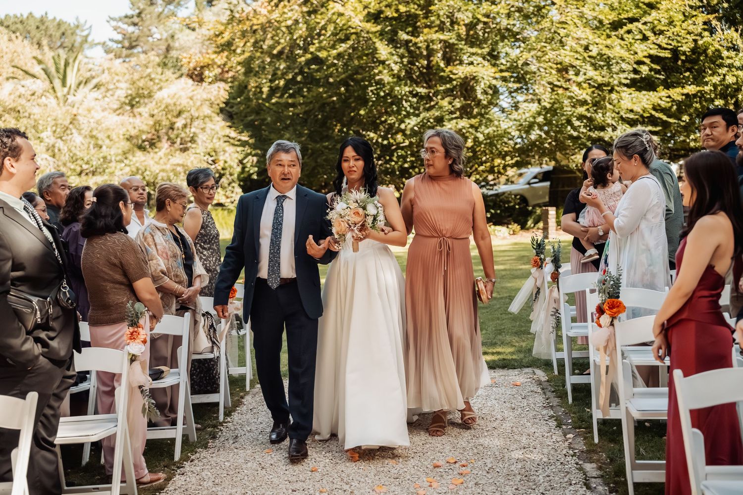 Bride walks down outdoor wedding aisle escorted by parents past white chairs and lush green trees in natural light.