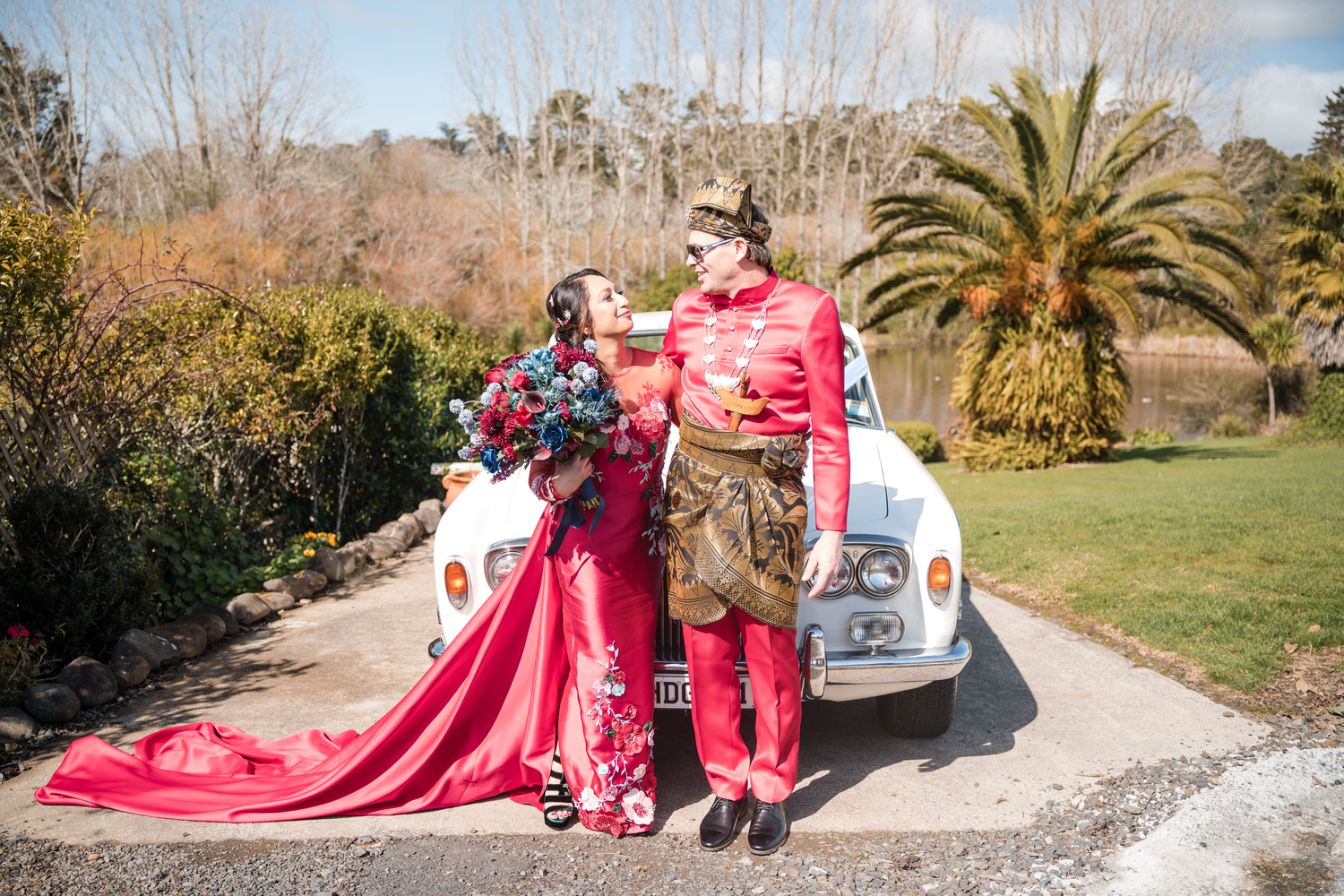 Couple in coordinated red outfits pose with their wedding car and trailing red dress.