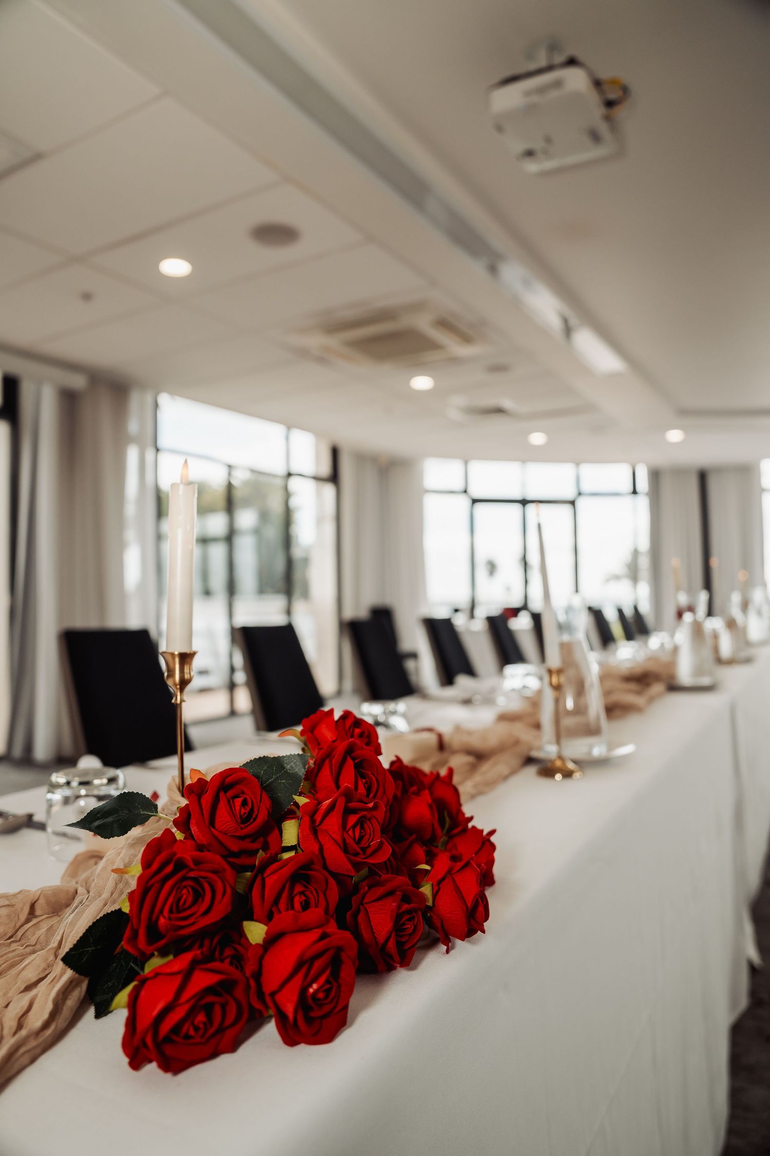 Red roses arranged on white tablecloths in an elegant indoor reception venue with modern lighting.