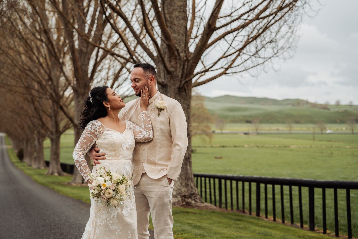 Wedding couple strolling along a scenic country path lined with bare trees and fencing.