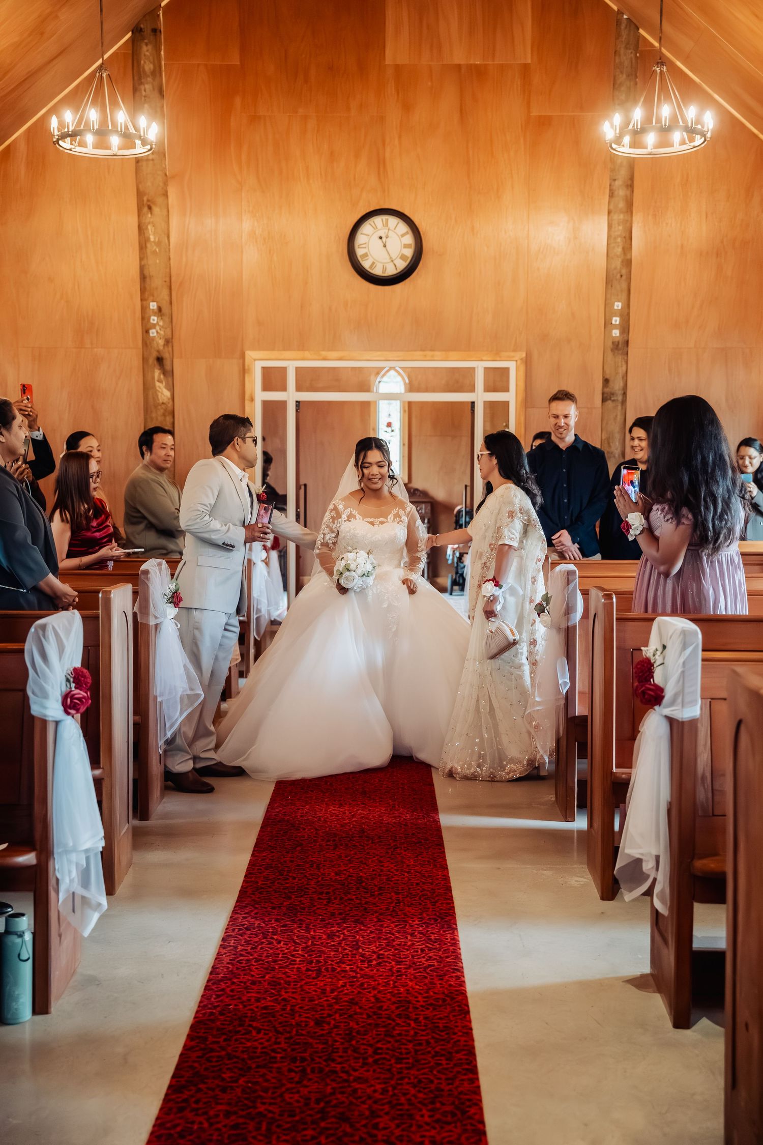 Bride walking down red carpet aisle in chapel wearing lace ball gown with guests standing.