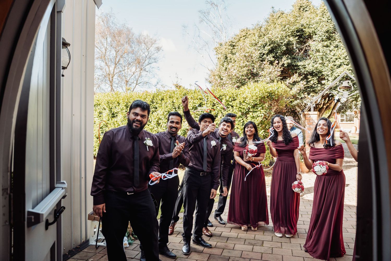 Wedding party in burgundy and black celebrating at doorway, bridesmaids and groomsmen cheering.