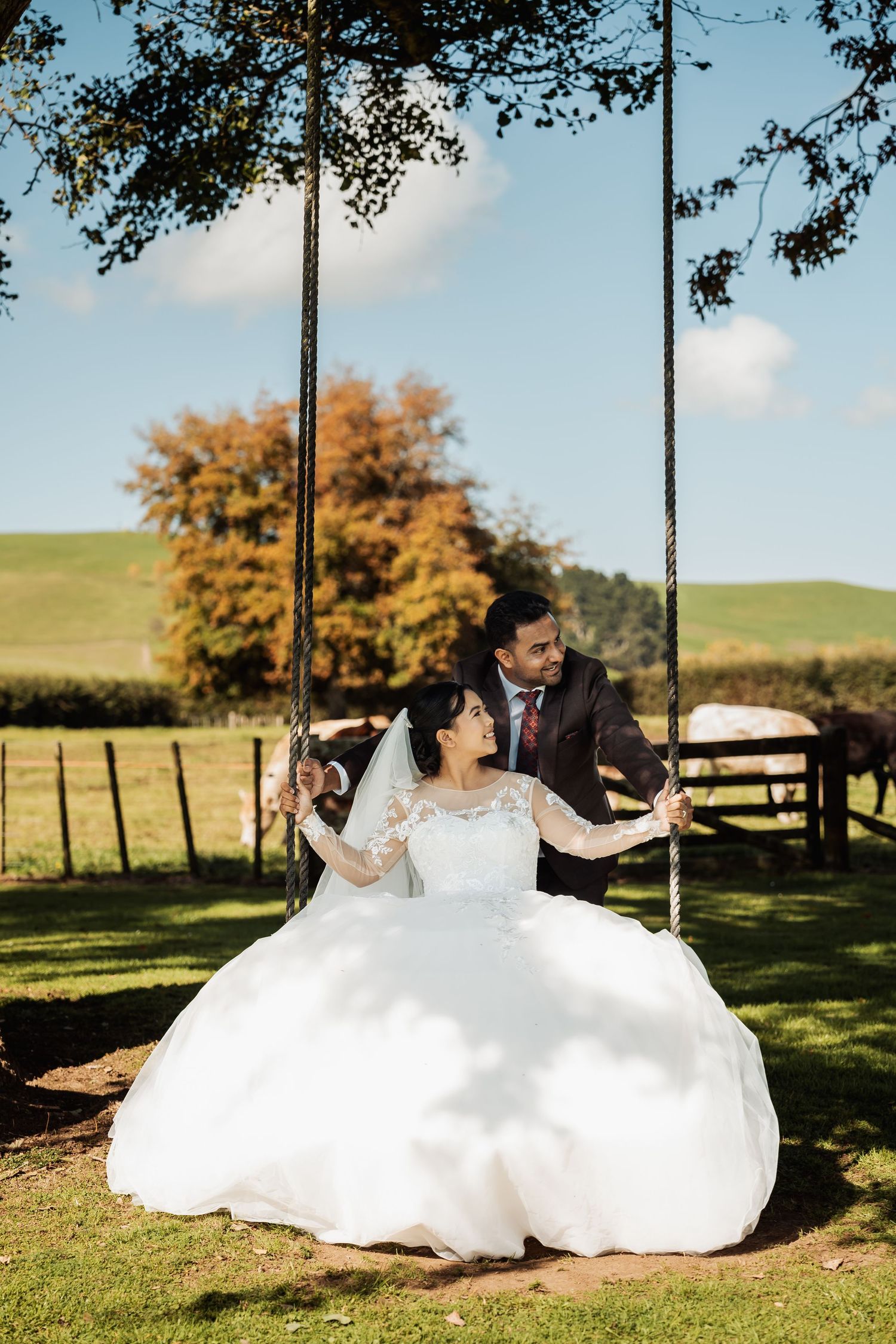 Bride sits on rope swing in white ball gown as groom stands behind her in autumn countryside.