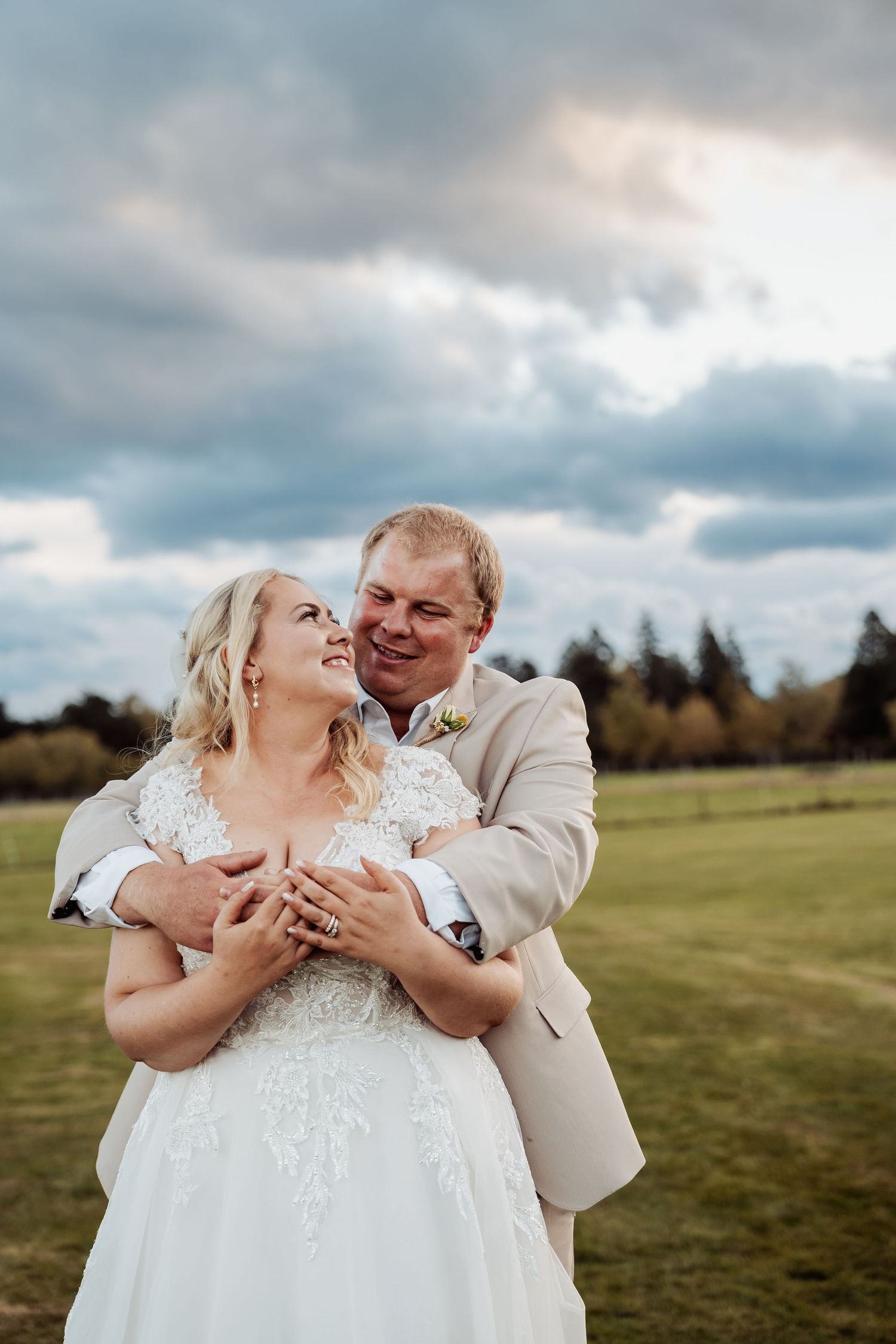 Smiling bride and groom embrace in open field under dramatic cloudy sky, groom in tan suit hugging bride from behind.