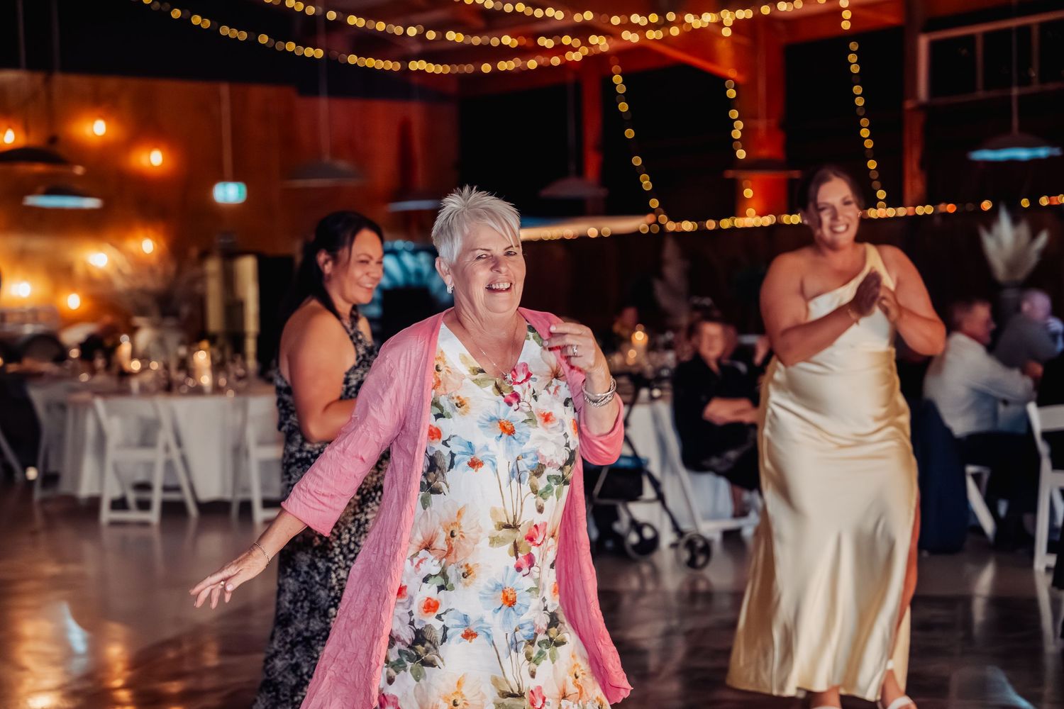 Elderly woman in floral dress and pink cardigan dances joyfully at a wedding reception under string lights.