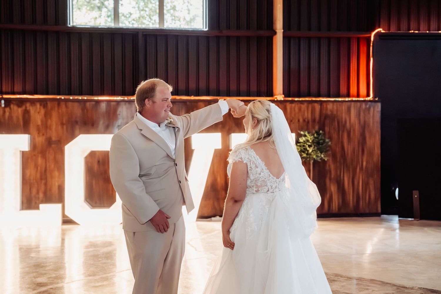 Groom lifting bride's veil during romantic wedding ceremony in rustic barn with glowing LOVE letter backdrop.