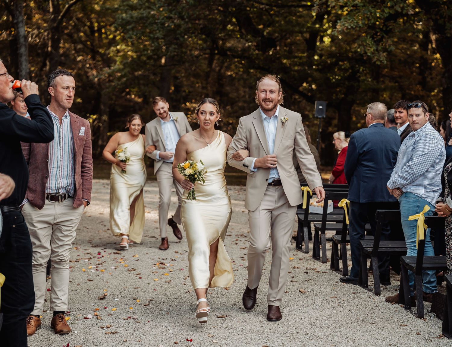Newlywed couple walking joyfully down outdoor aisle with wedding party following behind them.