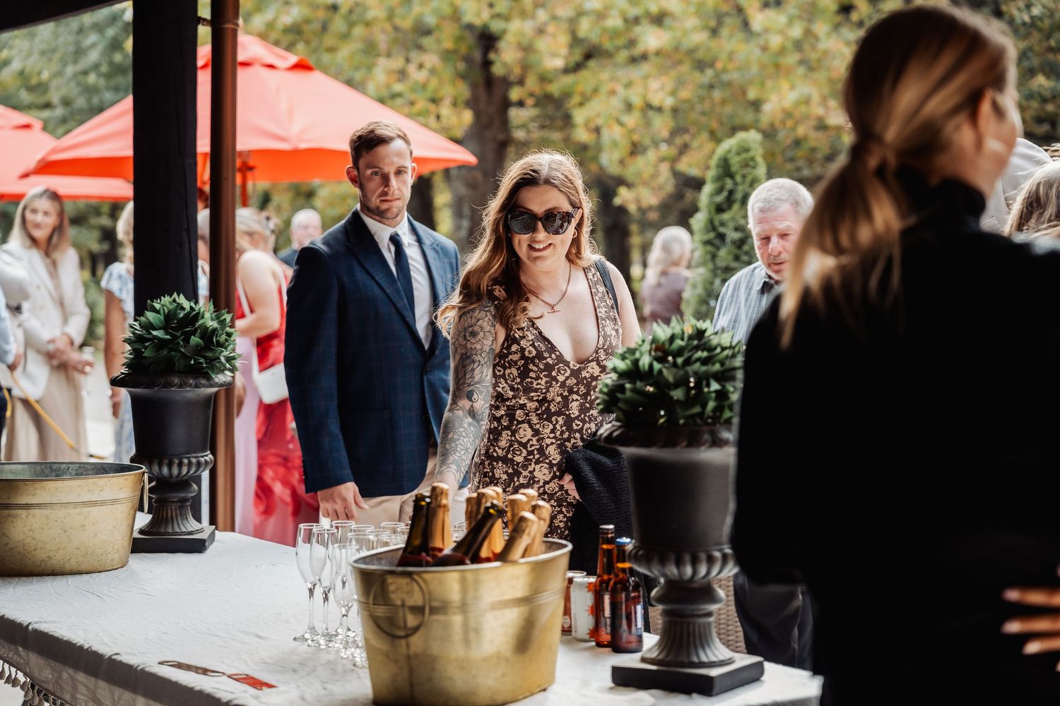 Wedding guests browse a drink station at an outdoor reception with red umbrella decor in the background.