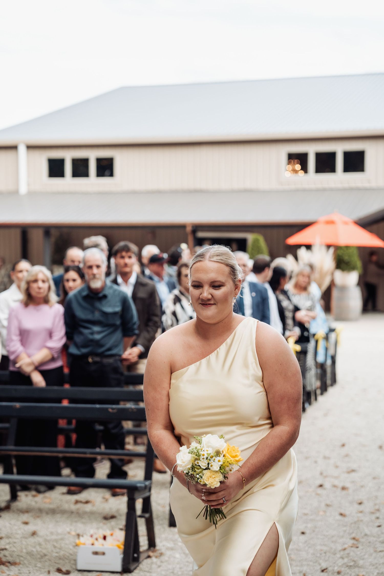 Bridesmaid in yellow one-shoulder gown walks down outdoor wedding aisle holding white and yellow bouquet.