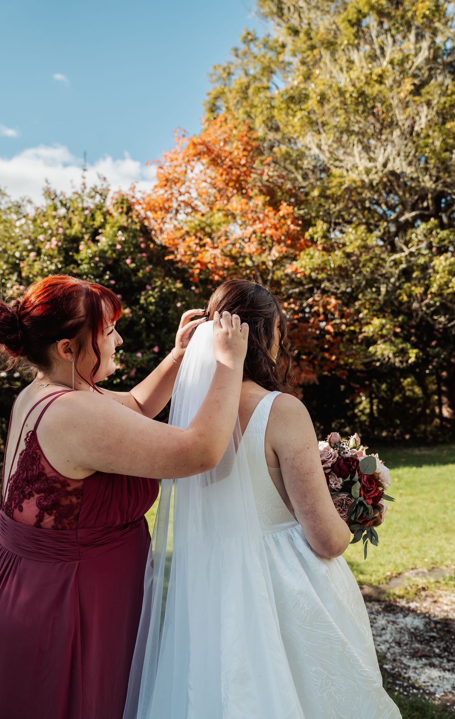 Bridesmaid in burgundy dress adjusting bride's white veil outdoors with autumn trees in background.