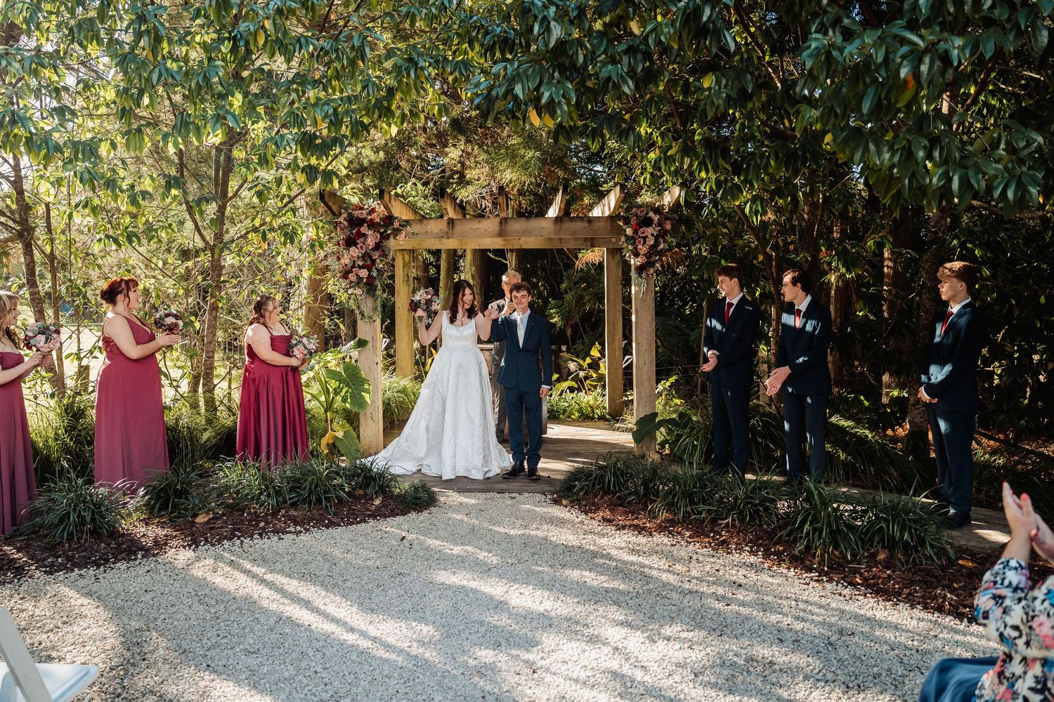 Bride and groom walk down aisle flanked by wedding party in garden ceremony.