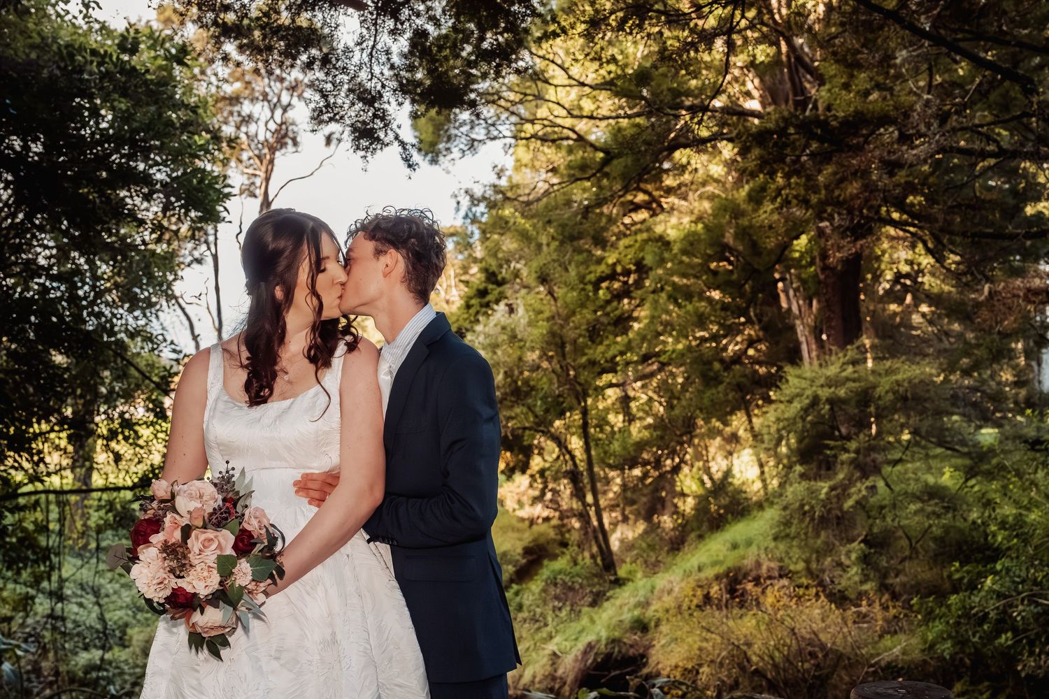 Bride and groom sharing a romantic kiss surrounded by lush green trees at their wedding.