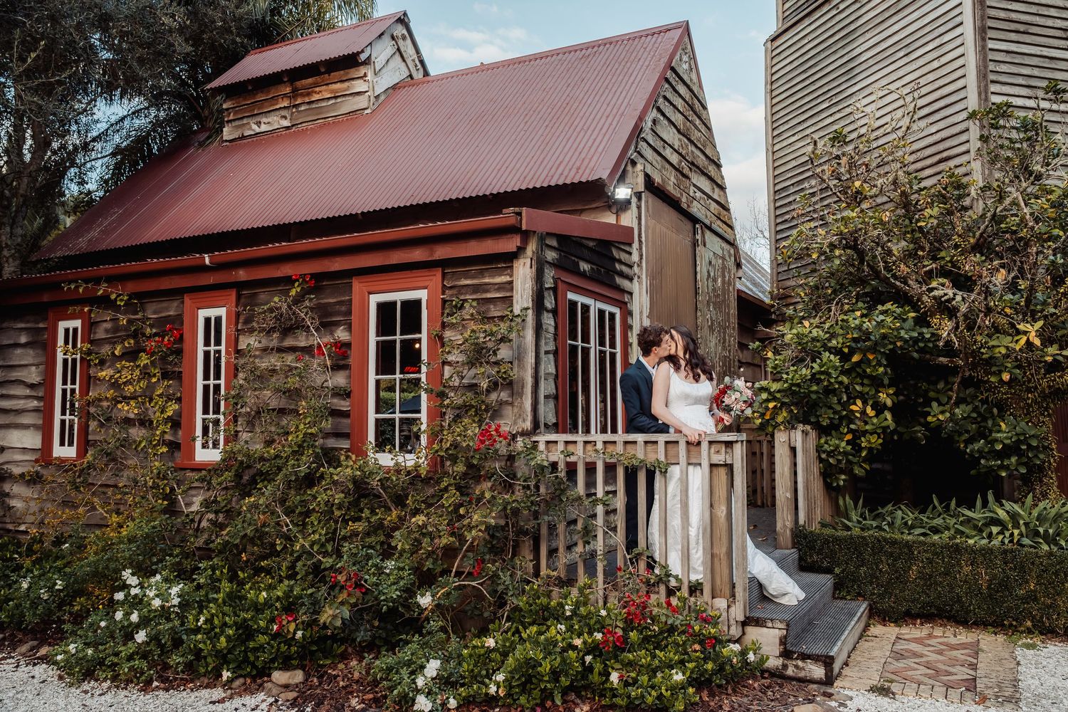 Bride and groom embrace on steps of rustic stone cottage with red roof and climbing roses.